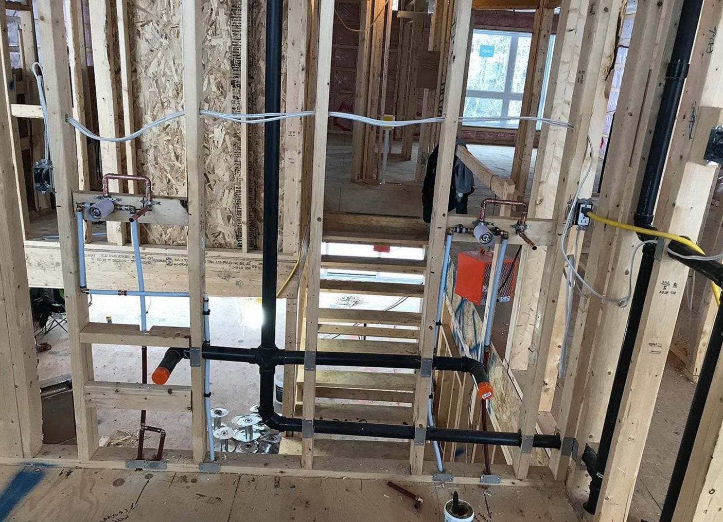 Open stud walls during a rough-in, with PEX supply lines, black ABS drain pipe and valve boxes set between the studs