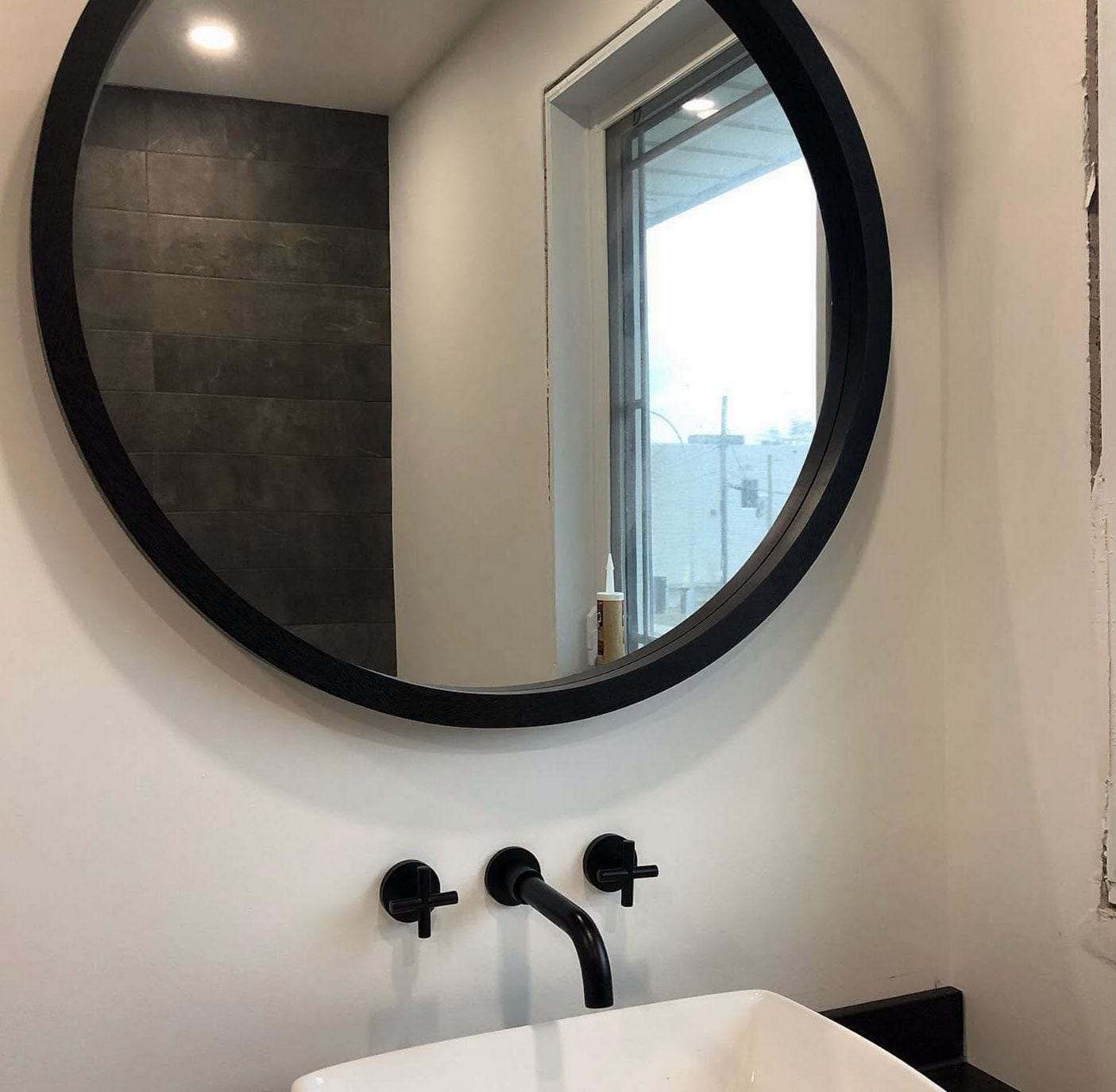 White vessel sink under a black two-handle wall-mount faucet and a round black-framed mirror