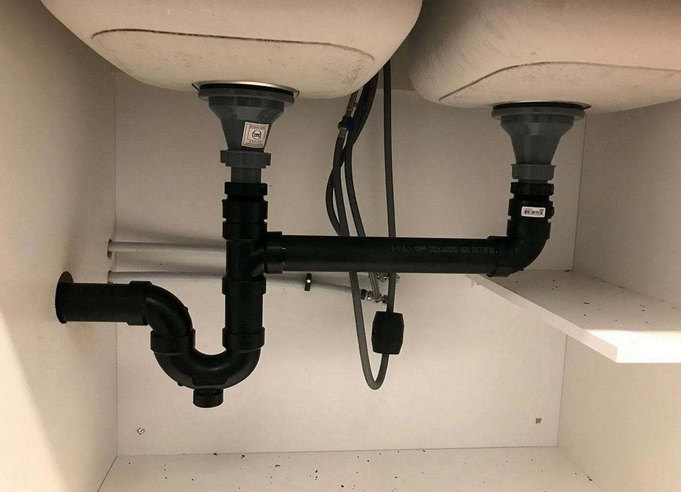 Double kitchen sink drain inside the cabinet: two basket strainers joined by black ABS pipe into one trap