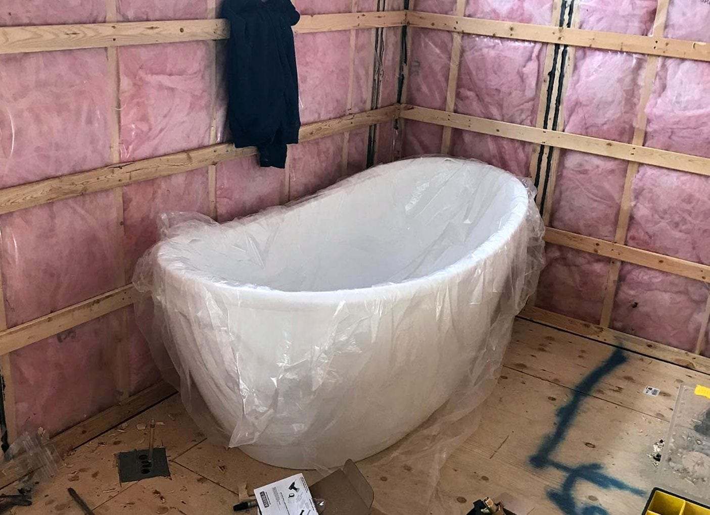 Freestanding white tub still in its plastic wrap in a bathroom under remodel, insulation showing between the studs
