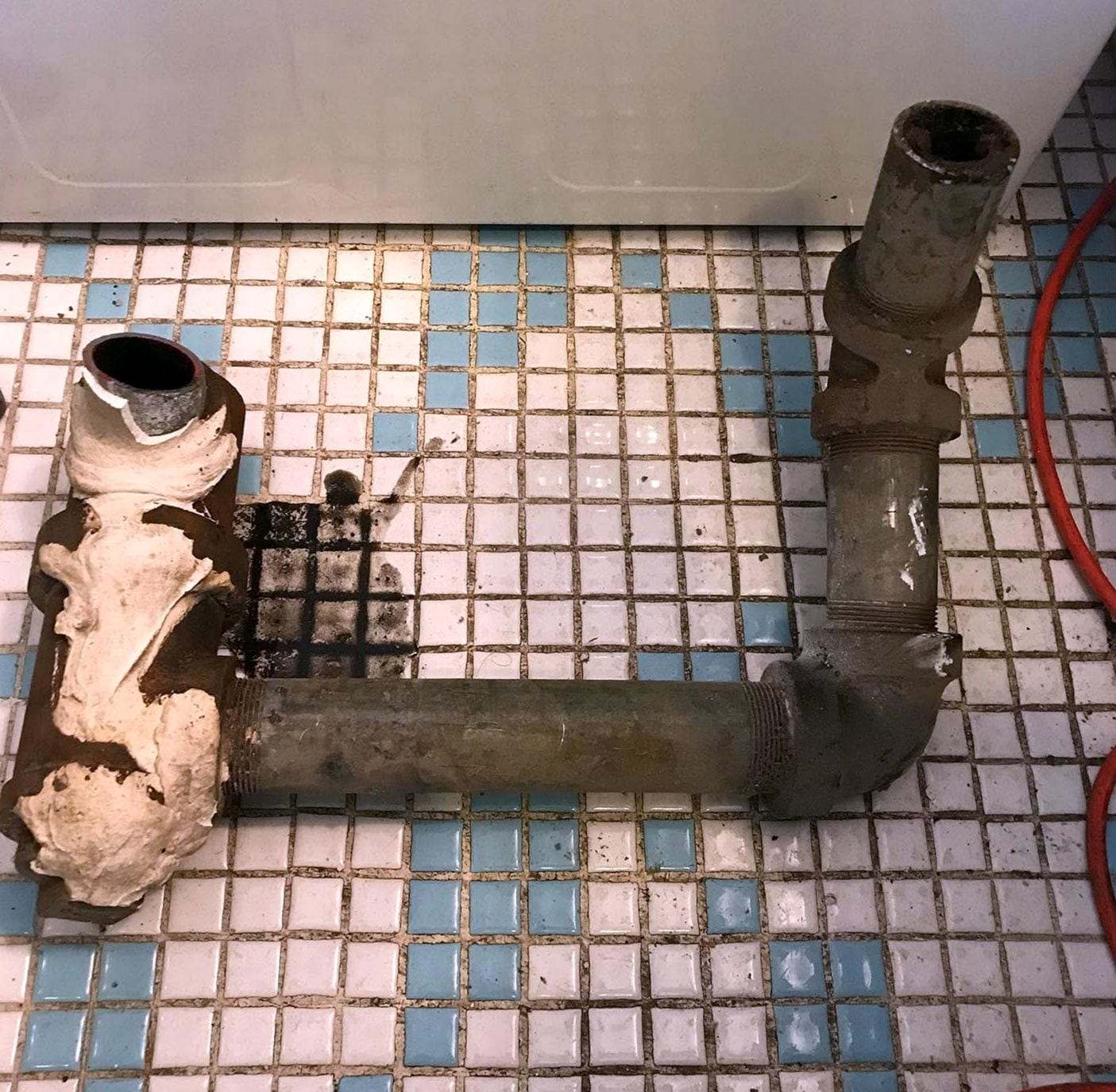 Old corroded drain pipe and elbow lying on a small-tile bathroom floor after it was cut out